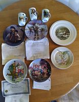 Overview photo of all collector plates and three mugs arranged on a wooden table, along with some certificates.