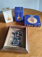 Photo showing two boxed M.J. Hummel Goebel bells, one boxed collector plate, and a framed print with children under umbrella on a wooden table
