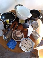 Top-down view of assorted kitchen items on wooden table showing various pots, colanders, utensils, and containers.