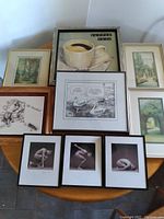 Seven framed artworks arranged on a wooden table; includes female nude photos, cartoons, landscapes, coffee painting
