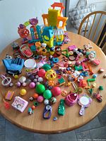 Top-down view of assorted colorful toys on a round wooden table, including a multi-level playset, dolls, balls, toy furniture, and various small accessories.