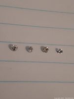 Four small round-cut diamonds placed horizontally on lined paper, showing size and detail.