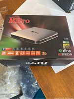 Photo of the RPro Android TV Box packaging showing product name, model image, and feature icons including Full HD 1080p, H.265, DLNA, EZMedia, 3G, USB, and various media format icons.