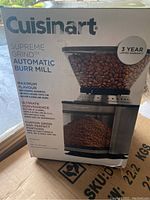 Box front showing Cuisinart Supreme Grind Automatic Burr Mill coffee grinder with beans and ground coffee, highlighting features such as 3 year warranty, maximum flavor, ultimate convenience, custom grind settings, and automatic shut-off timer.