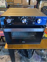 Front view of the Oster toaster oven showing control panel with three knobs, black and stainless steel exterior with dent on top.