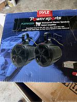 Pair of black cylindrical waterproof marine speakers with mounting clamps on top of the product box.