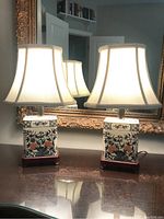Two matching ceramic table lamps on dark wood stands with off-white fabric shades placed on a wooden table in front of an ornate gold-framed mirror.