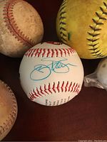 Close-up of signed baseball with blue ink showing signature and stitching, used baseball and softball in background.