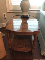 Full view of burled wood end table showing glass top with lamp and caned shelf below.