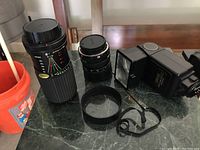 Photo showing large telephoto lens, smaller lens, black detachable lens hood, lens strap, and external flash unit grouped on marble table.