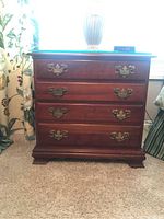 Front view of wooden dresser showing four drawers with ornate brass handles and glass top with lamp and alarm clock on top.