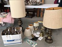 Two vintage table lamps with cream fabric shades, brass chandelier in box, small brass candlestick holders, additional lampshades, electrical components on garage floor