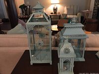 Front view of three turquoise decorative metal lanterns with glass panels showing detailed metalwork designs and bird figure on smallest lantern.