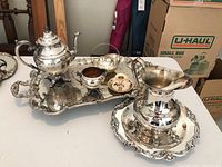 Silver plated serving dishes including teapot, sugar bowl, tray, wax candle holder, creamer pitcher, and matching tray arranged on table.