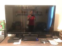 Full front view of Magnavox flat-screen TV placed on a wooden TV stand, remote controls and other electronic devices are placed on the stand surface.