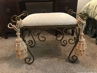 Front view of wrought iron vanity stool showing cushion, frame with scroll details and tassels.