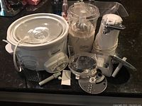 Photo of various kitchen appliances on a black countertop. Includes a Rival crockpot slow cooker, plastic lemonade pitcher, electric can opener, and assorted parts.