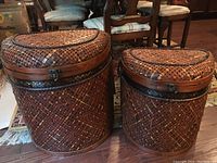 View of two woven wicker baskets with lids and black metal decorative bands placed on hardwood floor with dining chairs in background.