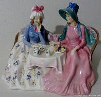 Porcelain figurine of two women seated at a tiny table set with teapot, cups, and small food items. One woman in pink dress and blue ribboned hat, the other with bonnet and blue jacket, floral skirt.