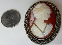 Close-up of cameo brooch with US quarter for size comparison, showing profile of woman in relief on reddish background with gold tone lattice bezel and rhinestone necklace pendant.