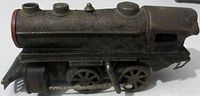 Side view of the pressed steel wind-up toy train engine showing the metal body, drive wheels, and wind-up key mechanism.