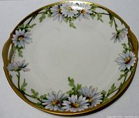 Front view of hand painted serving plate showing detailed daisy flowers with green leaves around the rim and gold accent trim and handles.