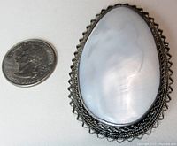 Top down photo showing the size of the brooch/pendant next to a US quarter. Teardrop shape, white iridescent Mother of Pearl shell, framed in detailed silver bezel.
