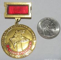 Front view of gold-tone circular Soviet WWII medal featuring a soldier carrying a wounded comrade, with red enamel banner and rectangular red enamel attachment bar, next to a US quarter for size.