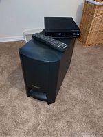 Photo of Bose Cinemate digital home theater subwoofer speaker and Magnavox DVD player with a remote control placed on top, on carpet floor.