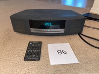 BOSE Wave Music System placed on carpeted floor with digital time display showing 12:03, accompanied by its remote and a paper marked '86'.