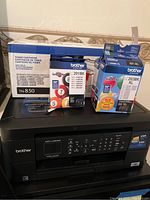 Brother MFC-J480DW printer with toner and ink cartridges displayed on top, showing product boxes and printer facade.