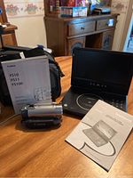 Photo showing Canon FS100 camcorder, black Canon carrying case, instruction manuals, and Magnavox portable DVD player with screen in open position on wooden table.