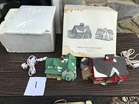 Two Dept 56 New England Farm Christmas village house figurines with original box, instructions and light cords visible. Both buildings show signs of wear; post broken on one house.