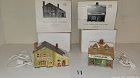 Two Dept 56 New England Village ceramic figurines in front of their boxes, with electrical cords attached for lighting.