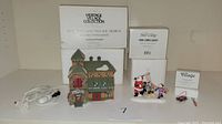 Overview photo showing all items: a village building with box and cord, 'Here Comes Santa' figurines, and sled & skis figurines with their boxes.