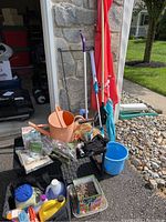 Image showing various gardening and outdoor items arranged by a garage. Included are a terracotta watering can, garden rake, spin scrubber mop, blue bucket, colorful floral pot, and other miscellaneous items.