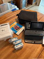 Photo showing printer, carrying case, instruction booklet, multiple packs of photo paper, and ink packs on a wooden table