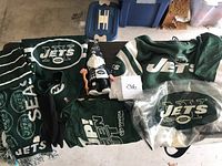 Multiple New York Jets fan items displayed on a table showing apparel, memorabilia and accessories including scarf, gloves, gnome figurine, rally towels, drawstring bag, and hooded sweatshirt.