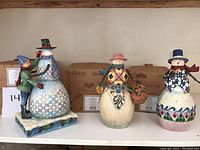 Front view of the three Jim Shore snowman figurines arranged in a row on a shelf, showing the boy with snowman, flower basket snowman, and pipe snowman.