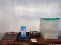 Photo of kitchen items including baking racks with red silicone handles, black metal dish holder, black baking pan, plastic 4 compartment storage box with green lid, and boxed spa socks.