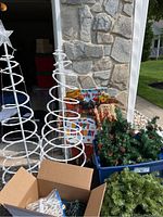 Two white metal spiral frame Christmas trees with star toppers, 'Happy Holidays' flag with a scarecrow figure, and blue plastic bin containing artificial greenery and pine cones, plus a cardboard box with string lights.