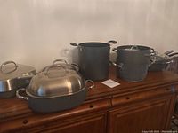 Overview showing 3 pots and several pans in black and metal finishes on wooden sideboard.