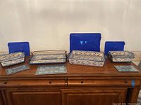 Full view of the four ceramic baking dishes with patterned designs with their respective blue plastic lids and three black metal wire carriers.
