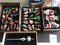 Top view showing three trays of glass Christmas ornaments in colorful designs including figurines of Santa Claus, birds, gifts, nutcrackers, and other festive shapes inside dark trays and a wooden crate with larger holiday figurines