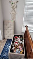 Beige tufted storage bench with interior filled with plush toys visible, macrame owl hanging above