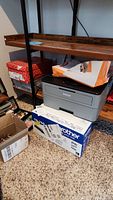 Gray Brother printer on carpet beneath wooden shelf, with toner cartridge box and pet food bag on top and nearby.