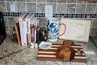 Cookbooks, ceramic bunny bookends, floral pitcher, cherry and maple striped cutting board, cat-themed cutting board and trinket dish, small salt and pepper bird shakers all arranged on a granite countertop in a kitchen setting.