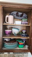 Cabinet shelf with pink mixing bowl, decorative bear bowl, various plates, plastic containers, and a pink electric kettle.