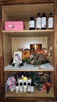Wooden shelf with several fragrance oil bottles, mini pink lock box, candle on floral arrangement, and bird decor on a frame pedestal