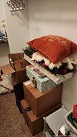 Stack of brown boxes with Christmas wreath, rust-colored decorative pillow, and miscellaneous items on top in a storage room.